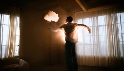 Person silhouetted against glowing cloud-shaped light suspended in a minimalist interior with large windows.