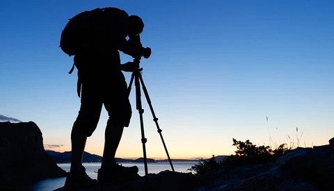 Silhouetted photographer on tripod against twilight sky and water.