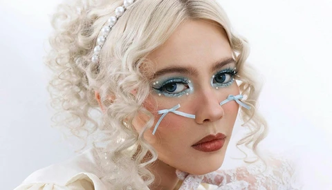 Portrait of a person with platinum blonde curly hair, dramatic eye makeup with blue eyeshadow, and decorative face accessories.
