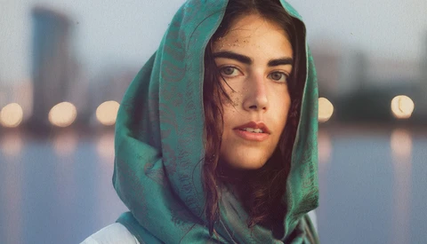 Woman wearing green headscarf looking at camera with urban waterfront and bokeh lights in background.