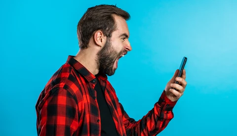 Bearded man in red plaid shirt holding smartphone against bright blue background.