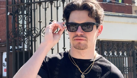Man wearing sunglasses and black textured shirt with gold chain necklace adjusting his glasses.