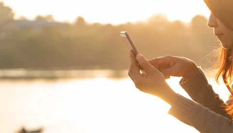 Woman holding smartphone toward water during golden hour, capturing landscape with backlighting.