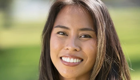 Woman with long dark hair smiling at camera outdoors with blurred green background.