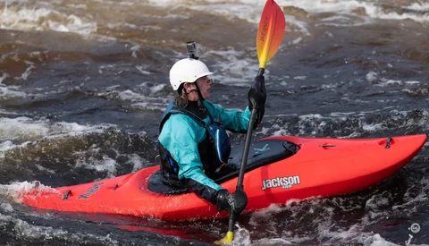 Whitewater kayaker in bright red kayak navigating turbulent rapids while holding yellow and red paddle.