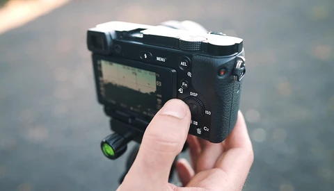 Hand holding a compact mirrorless camera with articulating screen and control dials visible.