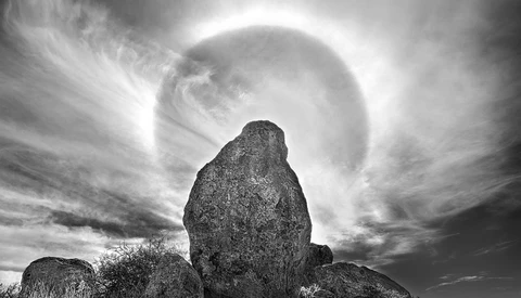 Large granite boulder silhouetted against a bright sun halo in dramatic black and white landscape.