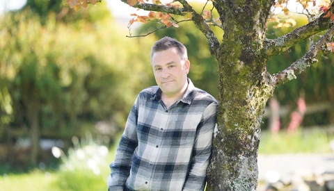 Man in blue plaid shirt leaning against a moss-covered tree trunk outdoors.