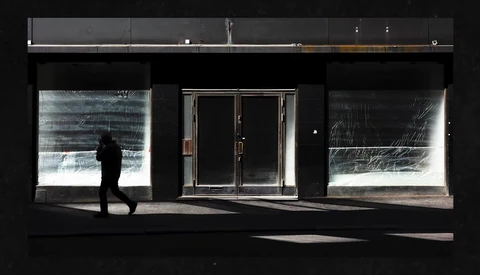 Silhouetted figure walking past storefront with illuminated window displays against dark facade.