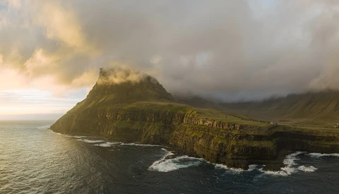 Dramatic coastal mountain with steep basalt cliffs rising from turbulent ocean waters under stormy skies.