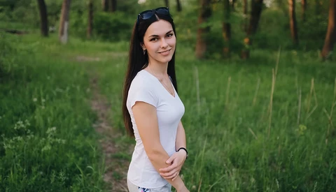 Woman in white t-shirt standing on a forest path with sunglasses on her head.
