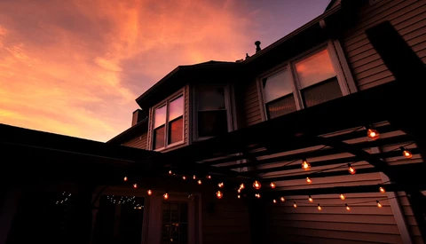 Two-story house silhouetted against vibrant orange and purple sunset sky, with warm string lights outlining the garage structure.