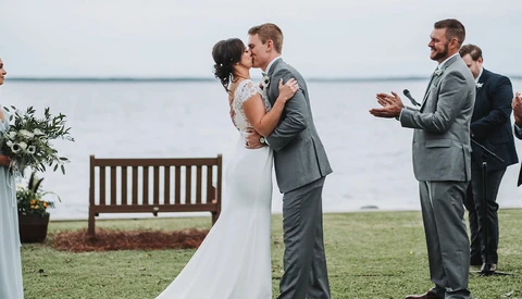 Groom and bride kiss during outdoor waterside ceremony as guests applaud.