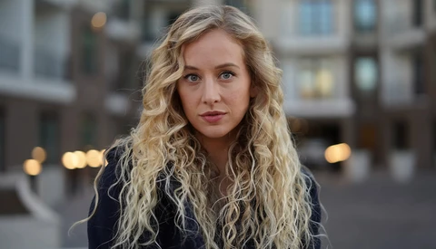 Woman with long blonde wavy hair looking at camera outdoors in urban setting.