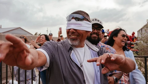 Man in white visor and sunglasses gestures toward camera while smiling at outdoor event with crowd in background.