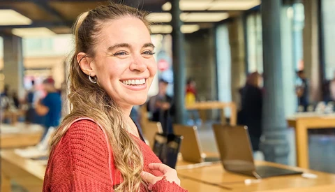 Young woman in red sweater smiling at camera in modern office space with wooden tables and blurred background activity.