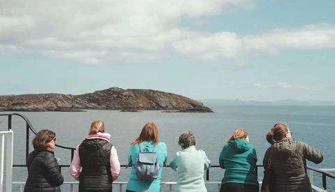 Six people viewing a rocky island from a boat railing on calm blue water.