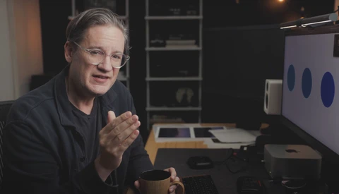 Man wearing glasses speaking at a desk with computer monitors and shelving in background.