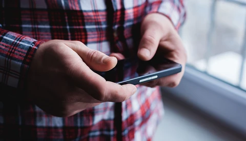 Close-up of hands holding a smartphone with a plaid shirt visible, shot near a window with natural lighting.