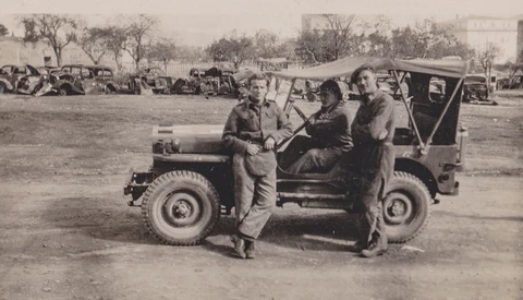 Three men in military uniforms standing beside a utility vehicle in an open field with vehicles and structures visible in the background.