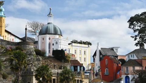 Hillside coastal town with white-domed chapel, yellow building, and colorful historic structures under partly cloudy sky.