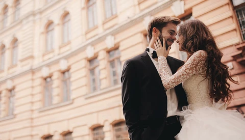 Bride and groom embrace in front of a classical peach-colored building facade.