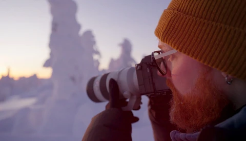Bearded photographer with orange beanie holding telephoto lens to eye during golden hour, snow-covered landscape blurred in background.