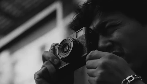 Photographer holding a film camera up to their face, captured in black and white from a close perspective.