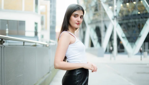 Young woman in white tank top and black leather skirt posing on urban pedestrian bridge.