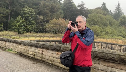 Male photographer in red and blue jacket holding a camera to his face while standing on a wooden boardwalk.