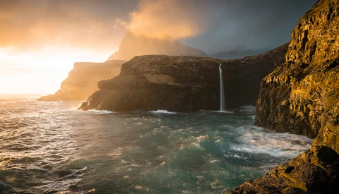 Waterfall cascading down golden cliffs into turquoise ocean waters during golden hour sunset.