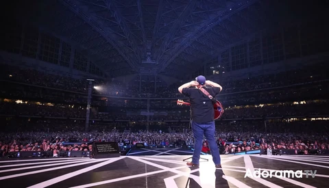 Performer on stage in large arena with dramatic blue overhead lighting and crowded audience.