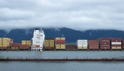 Large cargo ship stacked with colorful shipping containers against a mountainous coastline.