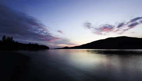 Twilight landscape with calm lake, forested shoreline, and mountains silhouetted against purple and pink sky.