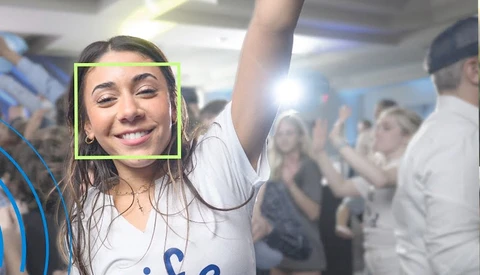 Smiling woman with arm raised at crowded indoor event, face detection box overlay visible.
