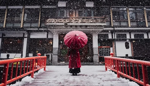 Person holding red umbrella walking across red bridge in heavy snowfall toward traditional architecture.