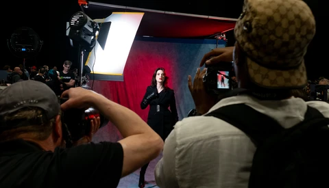 Audience members photograph a speaker on stage lit by professional studio lighting against a red and blue backdrop.