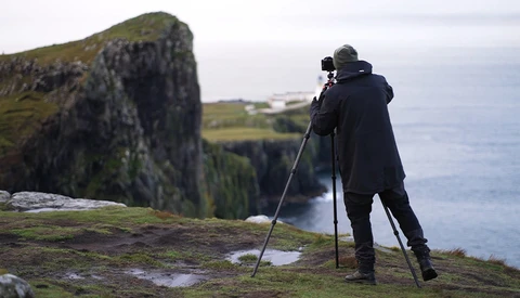 Photographer with tripod-mounted camera capturing coastal landscape from mossy cliff overlooking water.