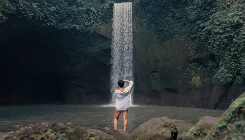 Person in white shirt standing in shallow water beneath a tall waterfall surrounded by lush vegetation.