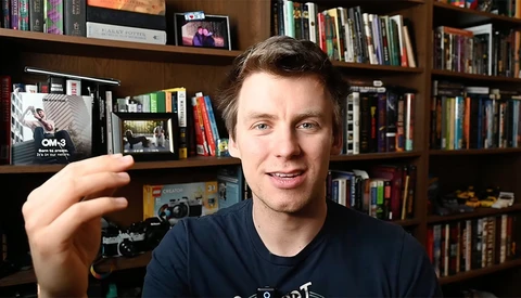 Photographer holding a compact camera while smiling at the camera in front of a wooden bookshelf.