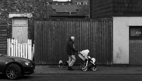 Person pushing a stroller across a street in front of a corrugated metal fence in black and white.