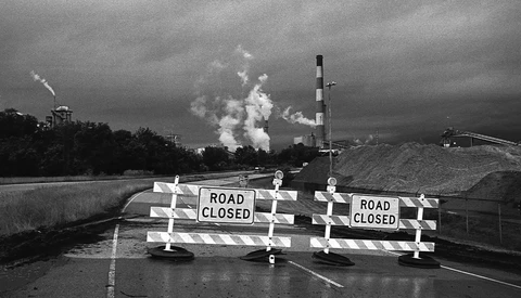 Road closed barricades blocking a highway with industrial smokestacks visible in the distance under dark skies.