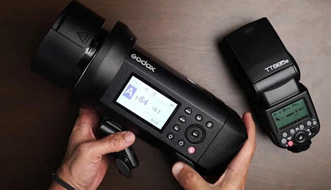 Hands holding a Godox studio flash with LCD screen and a Nikon TTL speedlight for comparison.