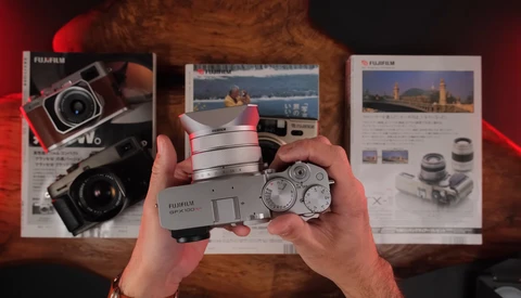 Hands holding a vintage compact rangefinder camera with product brochures and additional camera equipment displayed on a wooden surface.