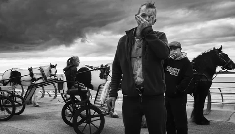 Man in dark coat and mask stands in foreground with horses and carts in dramatic black and white scene.
