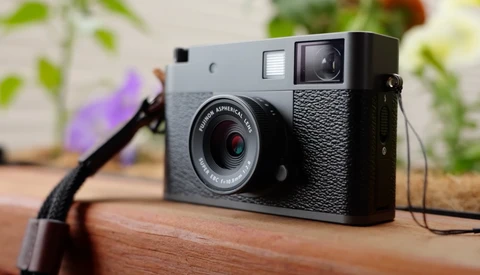 Vintage rangefinder film camera with gray and black body sitting on wooden surface with plants in background.