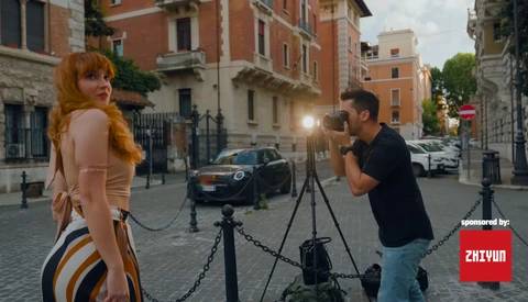Photographer on tripod shooting a female model posing on a European cobblestone street with historic brick buildings.