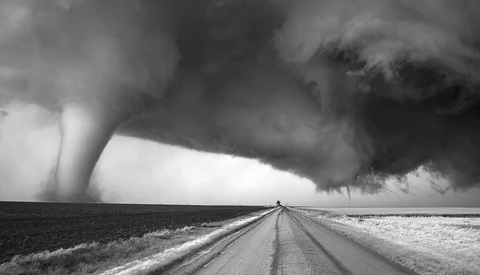Tornado touching down across flat plains with a straight road leading toward the storm under dark clouds.
