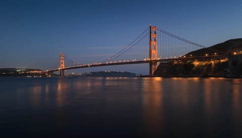 Suspension bridge illuminated at dusk, spanning a bay with calm water reflecting its lights.