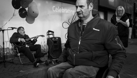 Man in black jacket sits on stage during an indoor event with musician and attendees visible in background.